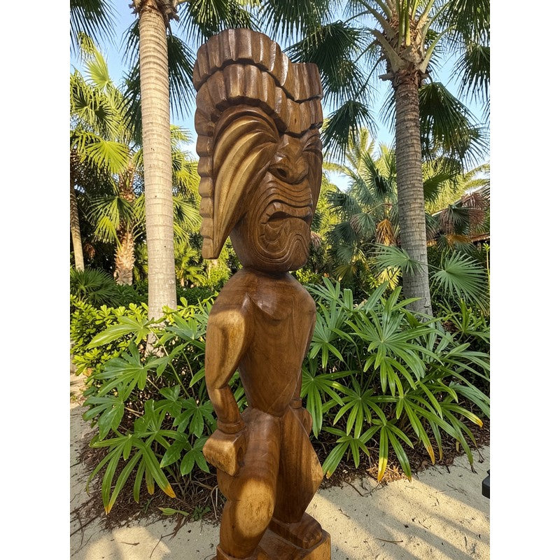 Wooden tiki statue in a tropical setting with palm trees and greenery.